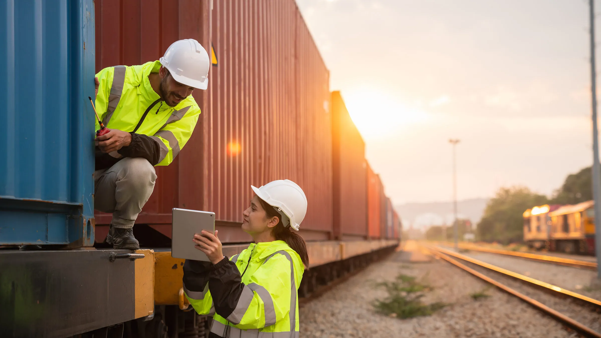 Zwei Mitarbeitende in Warnwesten prüfen im Abendlicht mit einem Tablet die Ladeeinheiten an einem Containerzug